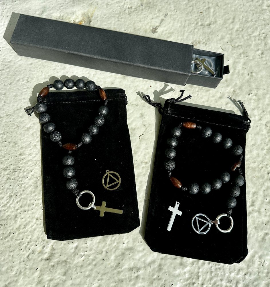 Two 12-Step Rosaries of black lava beads with wooden markers, laid on black velvet pouches in sunlight, each shown with a small cross and the AA circle-and-triangle pendant, beside a slim grey gift box.